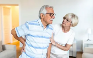 Older woman supporting her partner who is experiencing chronic back pain at home