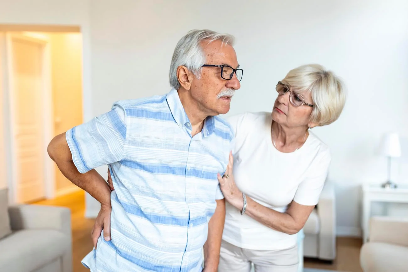Older woman supporting her partner who is experiencing chronic back pain at home